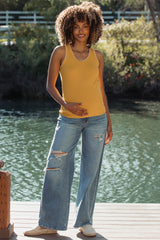 woman wearing a mustard yellow maternity tank top and distressed wide-leg jeans, holding her baby bump by the water