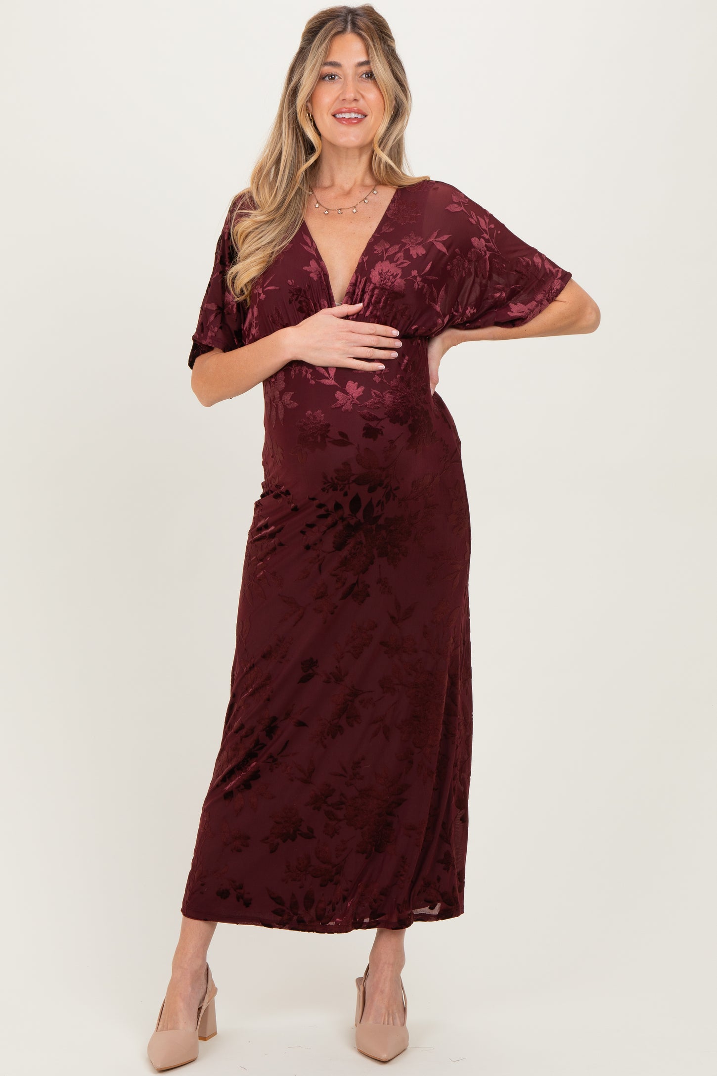 woman wearing a burgundy floral velvet maternity maxi dress with a deep v-neck and flutter sleeves while cradling her baby bump