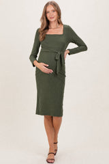 woman wearing a dark green ribbed maternity dress with a square neckline and tied waist, cradling her baby bump