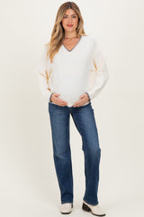 woman wearing a cream maternity sweater with a v-neck and blue trim, holding her baby bump while standing in jeans