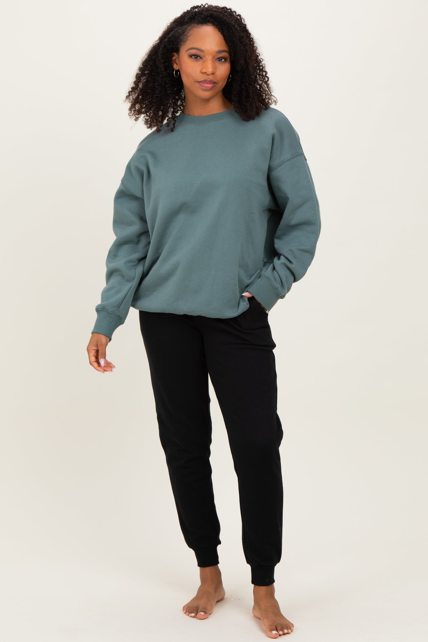 woman wearing a teal oversized sweatshirt and black jogger pants, standing barefoot against a neutral background