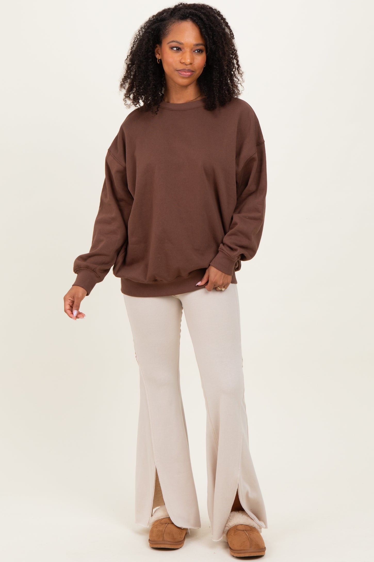 woman wearing a brown oversized sweatshirt and beige flare pants, standing in a relaxed pose