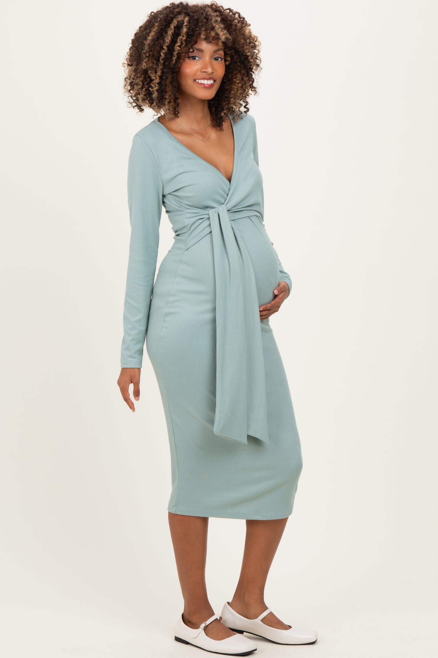 woman wearing a sage green long sleeve maternity dress with a tie at the waist and holding her baby bump