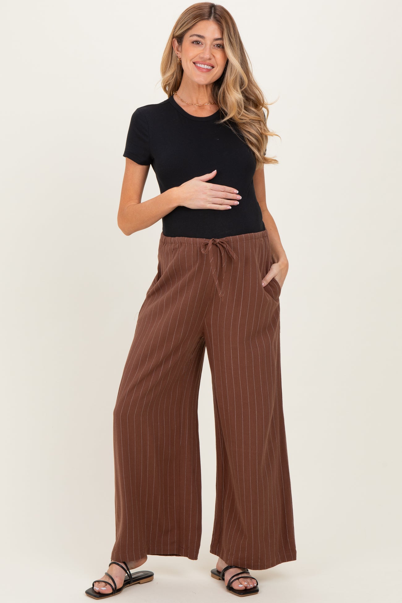 woman wearing a black short sleeve top and brown striped maternity wide-leg pants, holding her baby bump