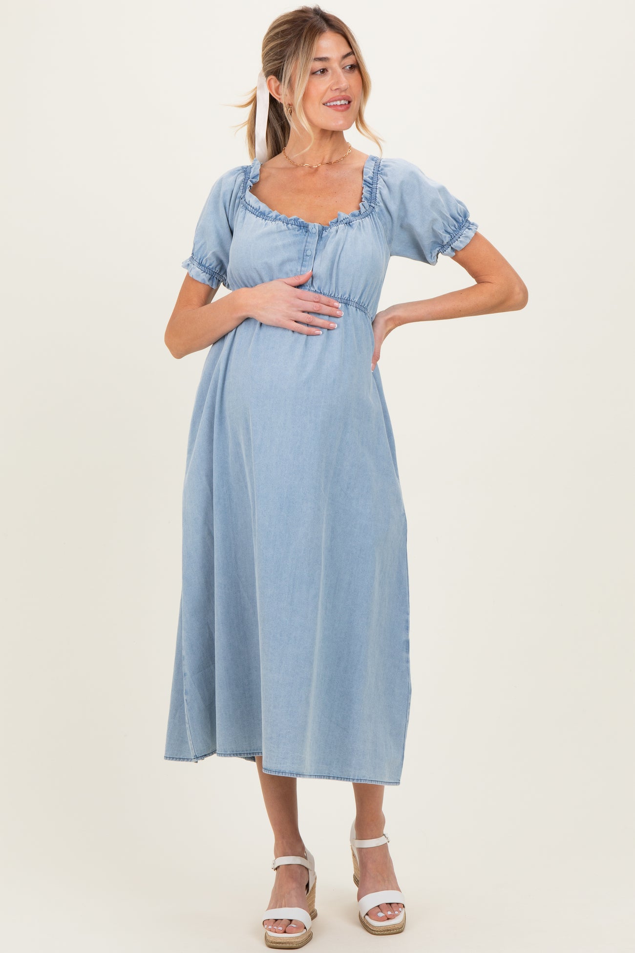 woman wearing a light blue denim maternity dress with short puff sleeves and a square neckline, holding her baby bump
