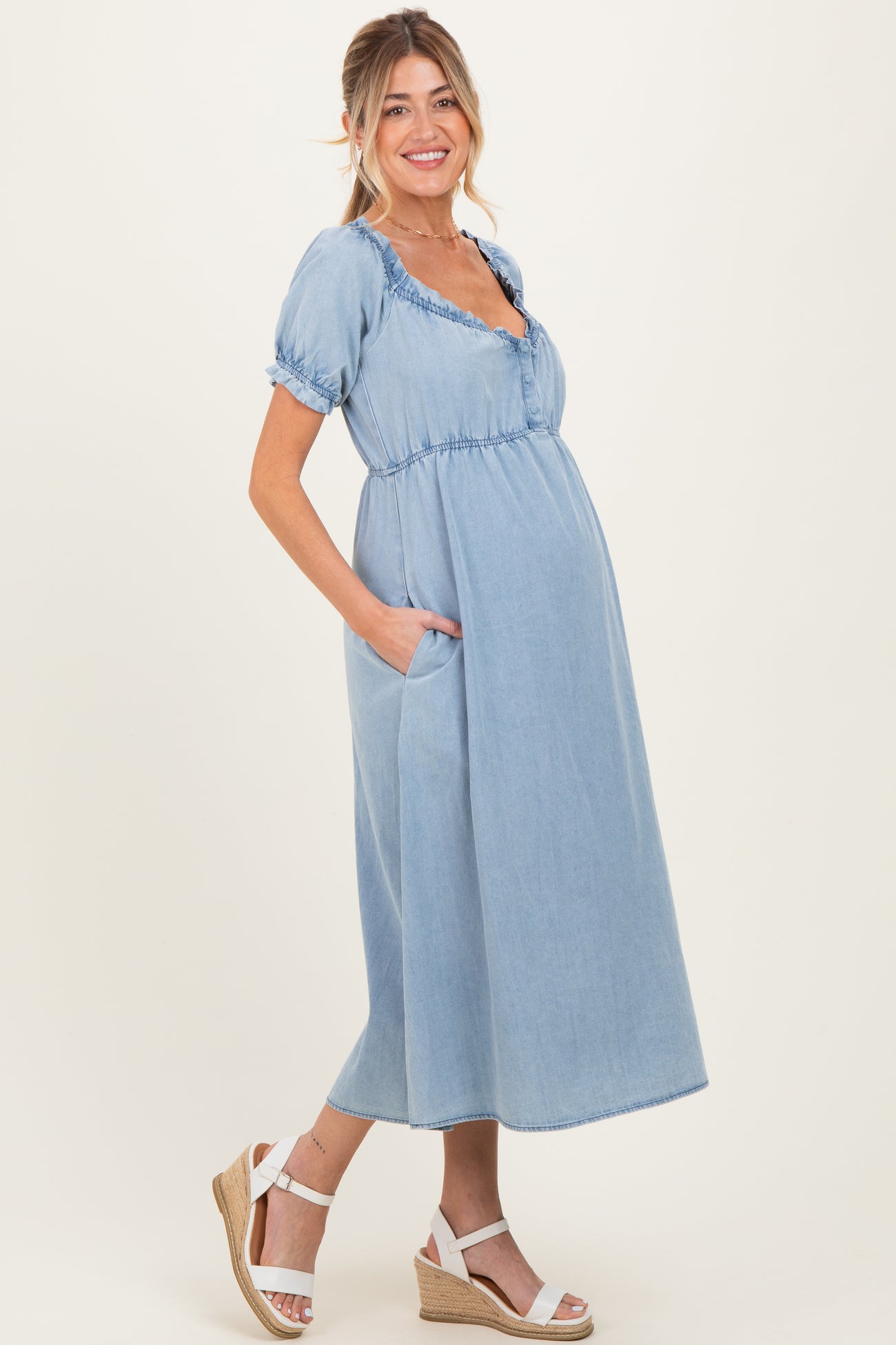 woman wearing a light blue denim maternity dress with short puff sleeves and pockets, showing her baby bump