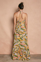 woman wearing a colorful patterned halter maxi dress with a tiered hem and open back, standing against a warm background