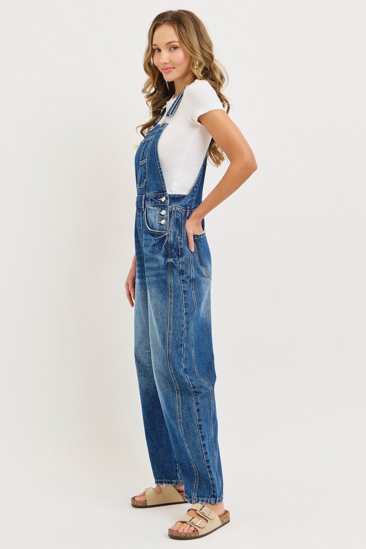 woman wearing a white short sleeve t-shirt and blue denim overalls, standing sideways with pockets visible