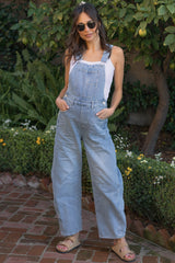 woman wearing a light blue maternity overall with wide legs and a white tank top, standing outdoors with sunglasses on