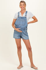 woman wearing a light blue denim maternity short overalls and a gray short sleeve top, holding her baby bump