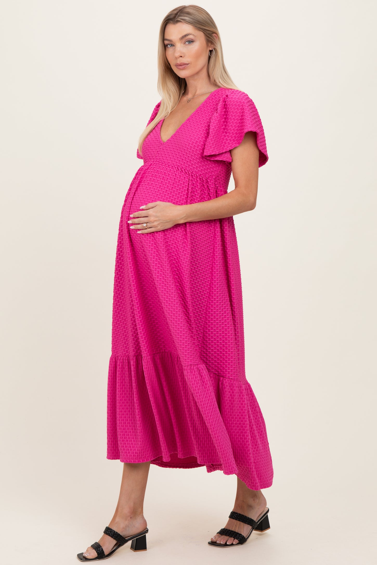 woman wearing a bright pink maternity dress with flutter sleeves and a tiered hem, gently cradling her baby bump