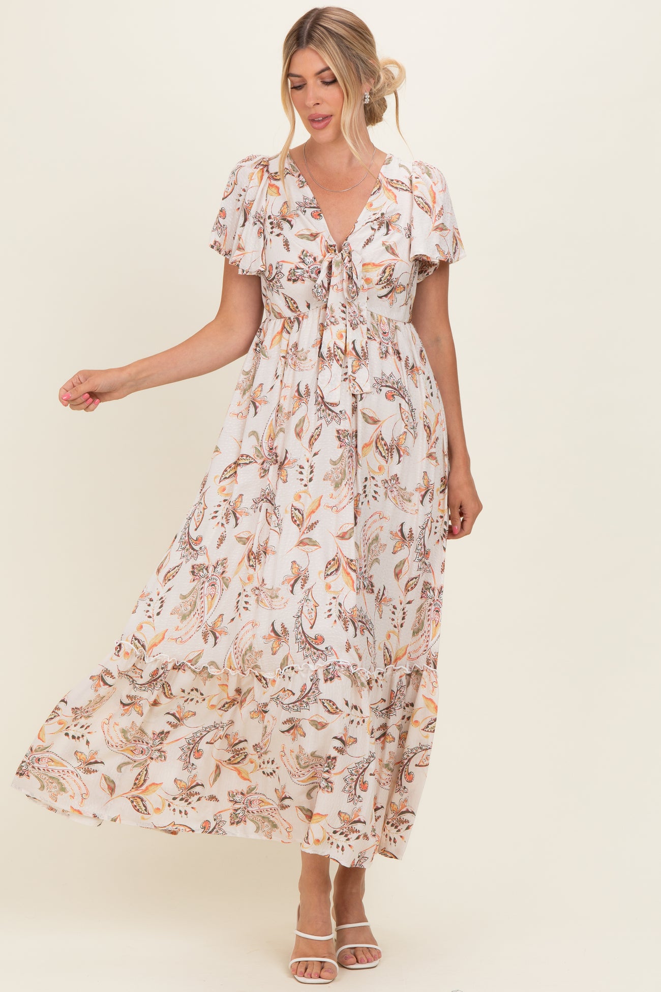 woman wearing a floral print maxi dress with short sleeves and a tie-front detail, standing with one hand raised