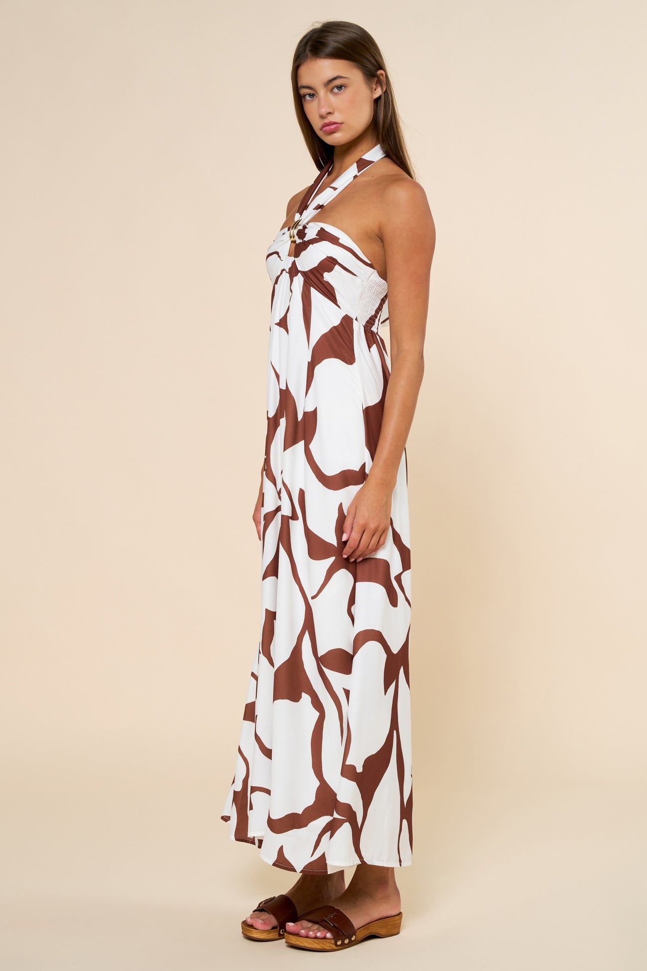 woman wearing a white maxi dress with brown abstract prints and a halter neck style, standing sideways