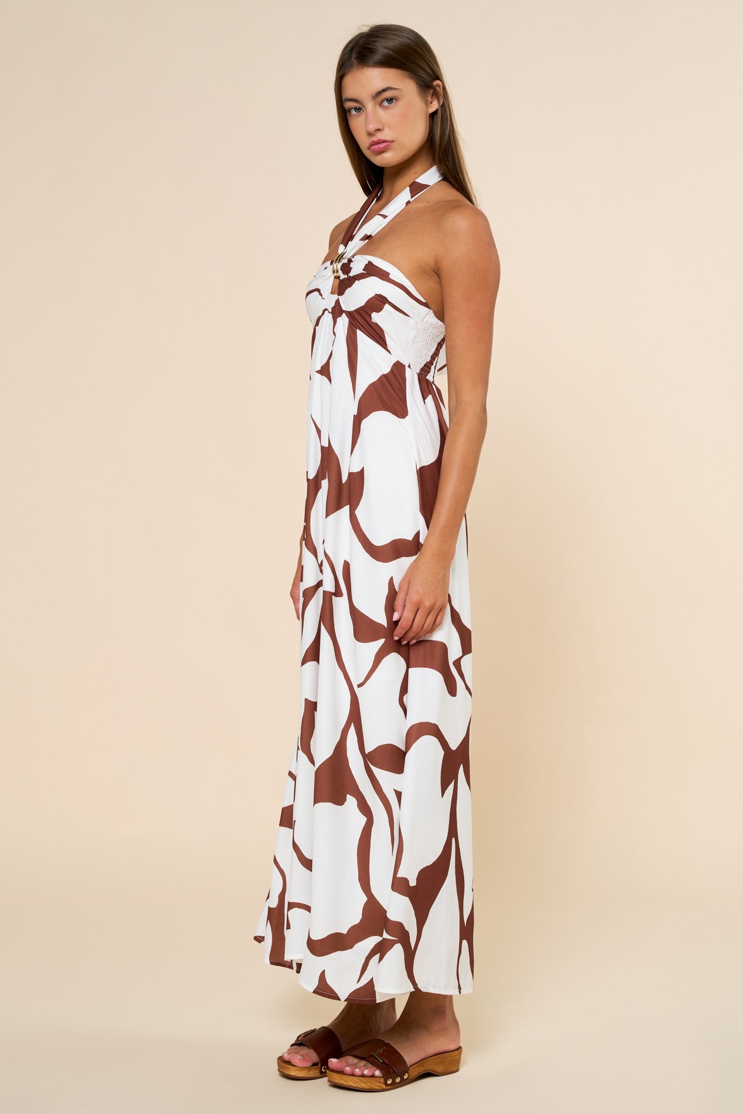woman wearing a white maxi dress with brown abstract prints and a halter neck style, standing sideways
