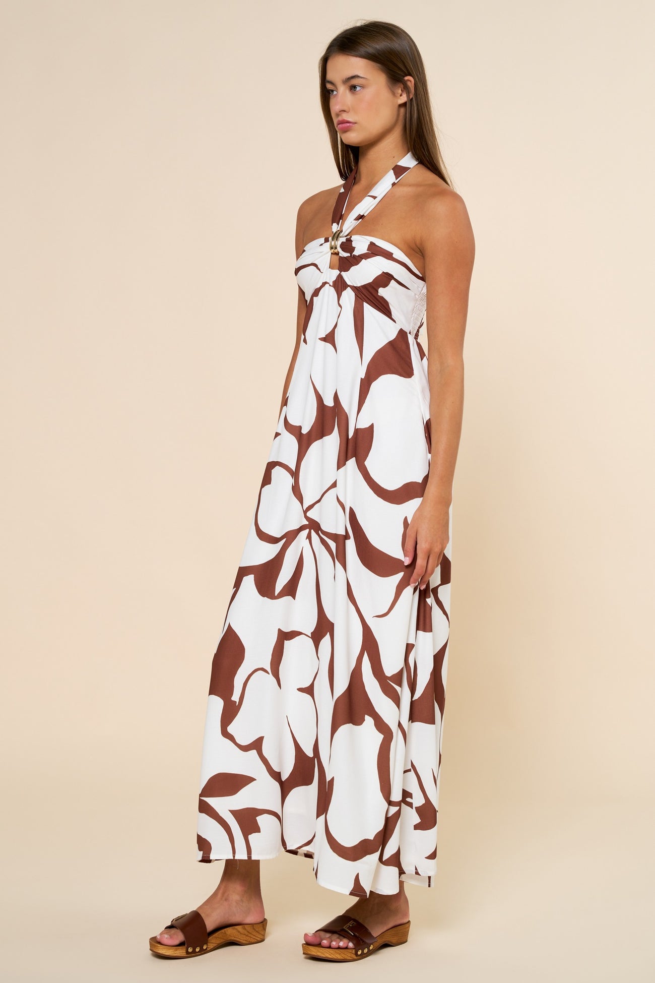 woman wearing a white maxi dress with brown floral print and a halter neck design, standing on a light background