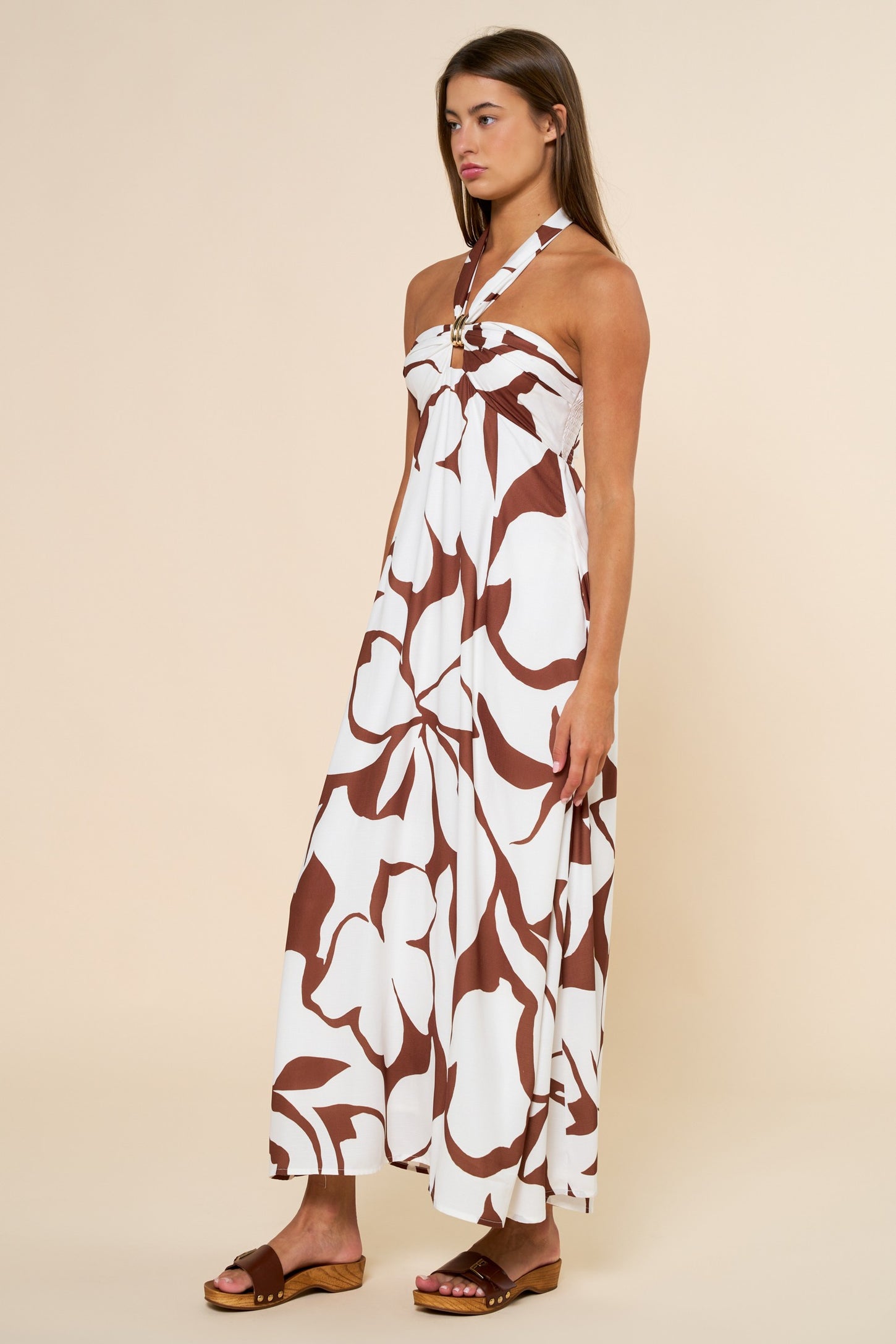 woman wearing a white maxi dress with brown floral print and a halter neck design, standing on a light background