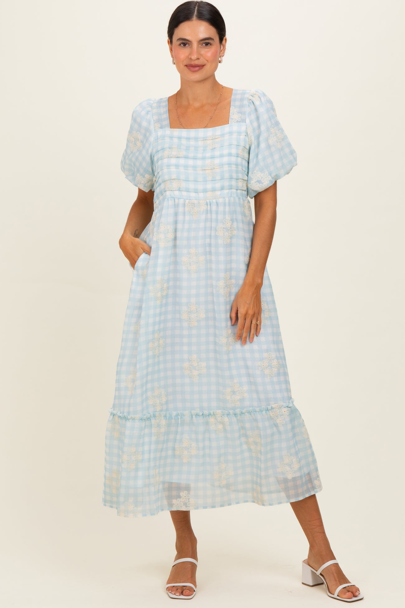 woman wearing a light blue gingham midi dress with puff sleeves and floral details, standing with hands in pockets