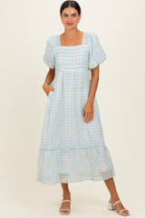 woman wearing a light blue gingham midi dress with puff sleeves and floral details, standing with hands in pockets