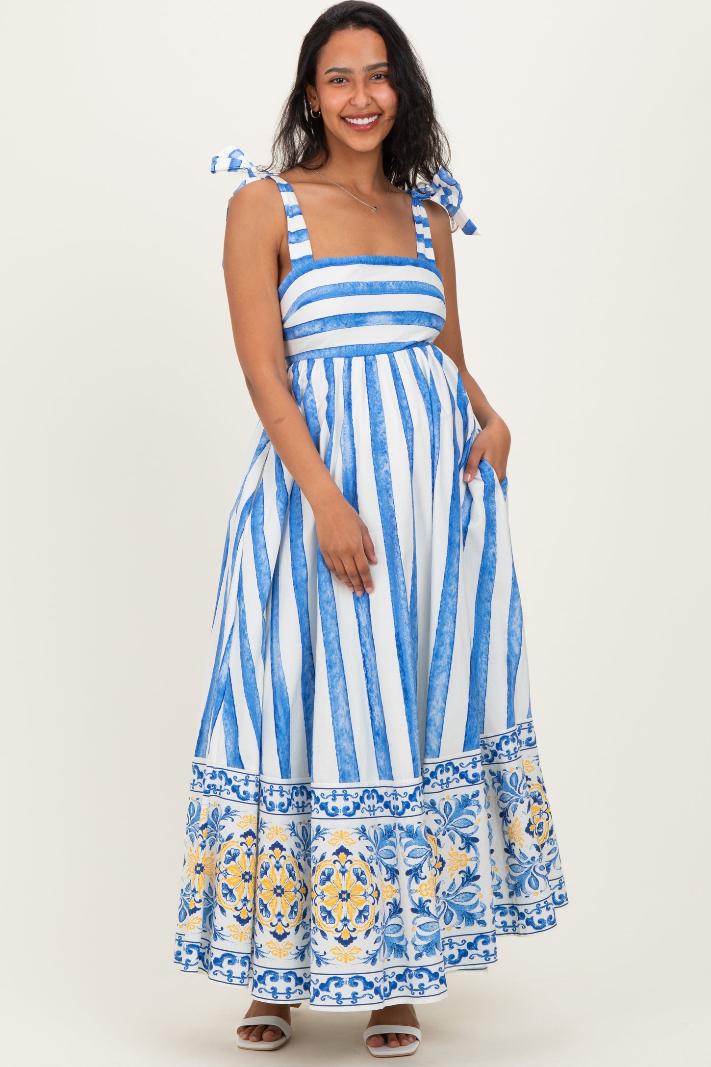 woman wearing a blue and white striped maternity maxi dress with tie straps and a floral print at the hem, holding her baby bump