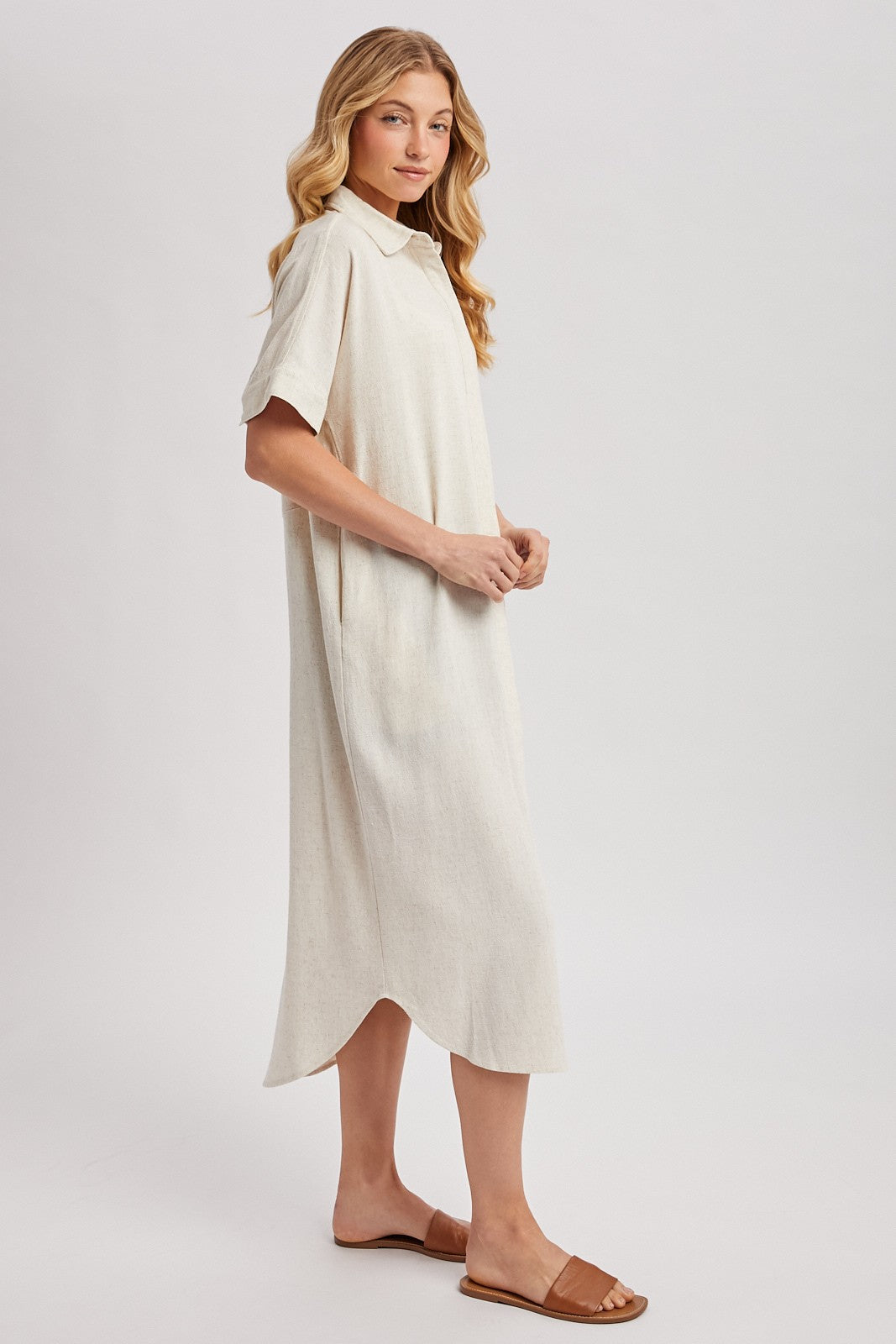 woman wearing a light beige short sleeve midi dress with a collared neckline and side slits, standing in profile