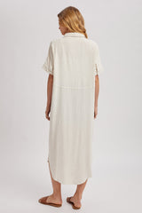 woman wearing a light beige short sleeve shirt dress with a back slit and relaxed fit, shown from the back