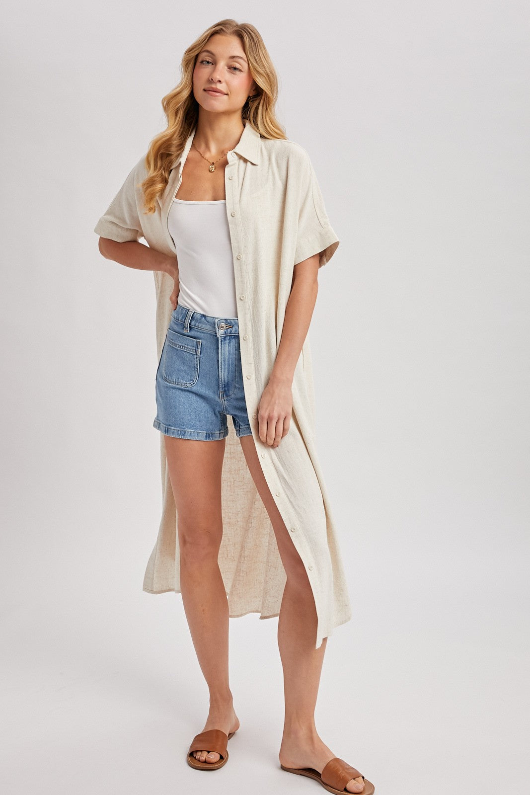 woman wearing a cream-colored button-up shirt dress over a white tank top and blue denim shorts with sandals