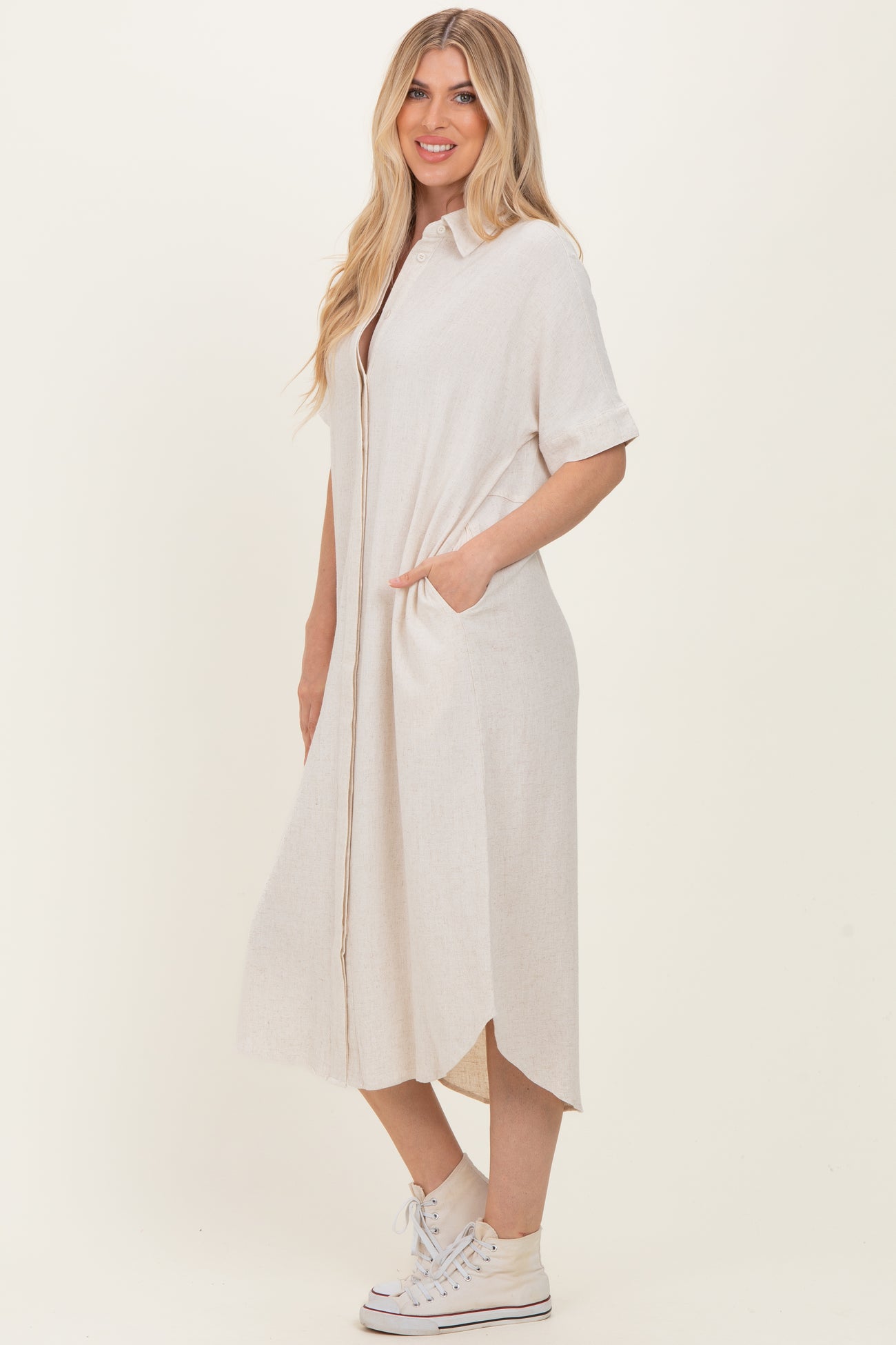 woman wearing a light beige short sleeve button-up dress with side pockets and a high-low hem, paired with white sneakers