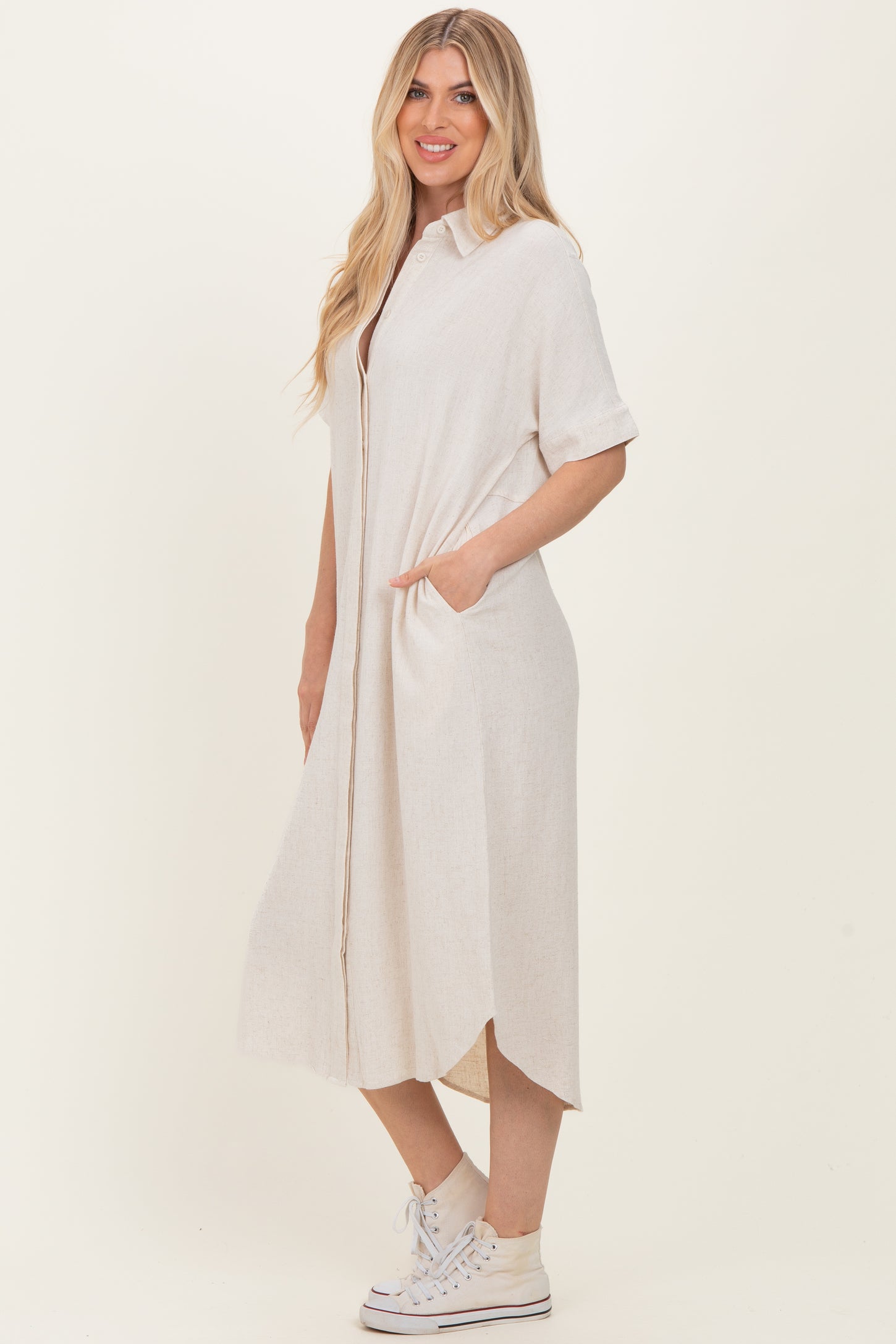 woman wearing a light beige short sleeve button-up dress with side pockets and a high-low hem, paired with white sneakers