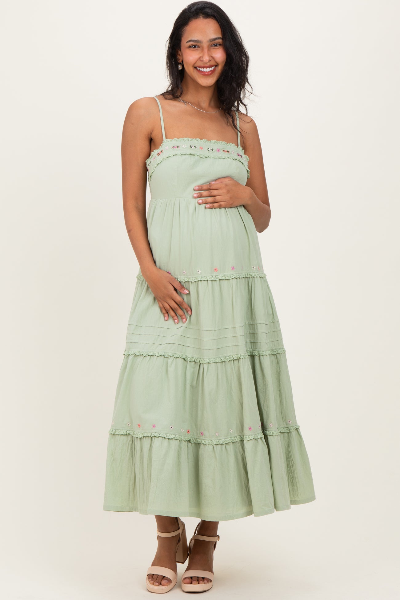 woman wearing a light green maternity dress with tiered design and floral embroidery, cradling her baby bump