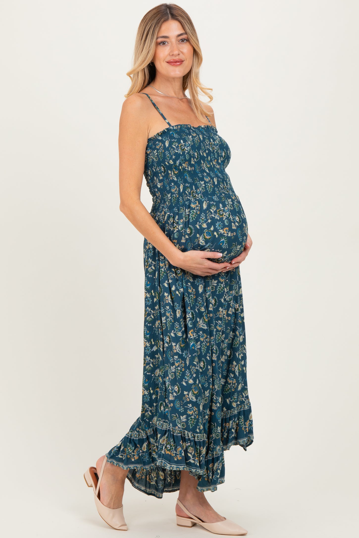 woman wearing a floral maxi maternity dress in teal, holding her baby bump with a smile