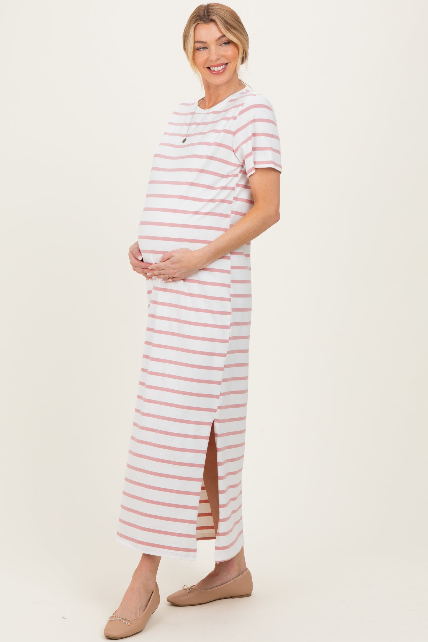 woman wearing a striped pink and white maternity maxi dress, holding her baby bump with side slits, paired with flat shoes