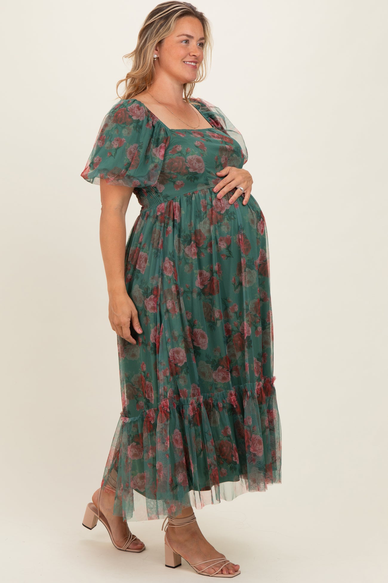 woman wearing a green floral maternity plus dress with puff sleeves and a ruffled hem, holding her baby bump