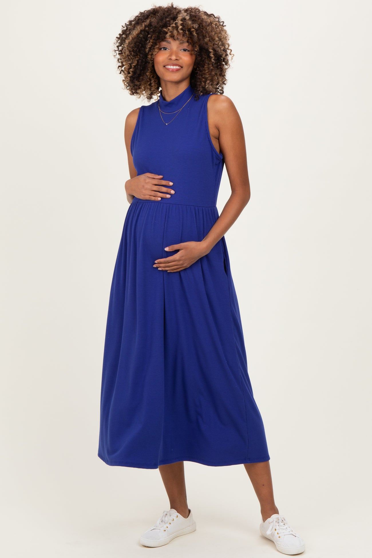 woman wearing a sleeveless blue maternity dress, cradling her baby bump and smiling