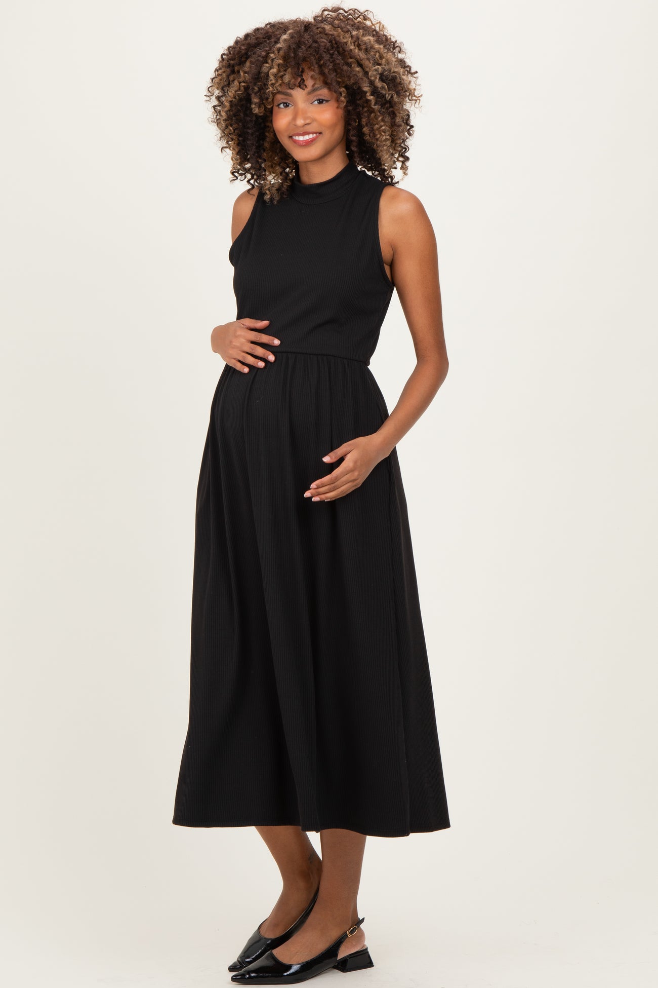 woman wearing a black sleeveless maternity dress with a fitted bodice and flowing skirt, cradling her baby bump