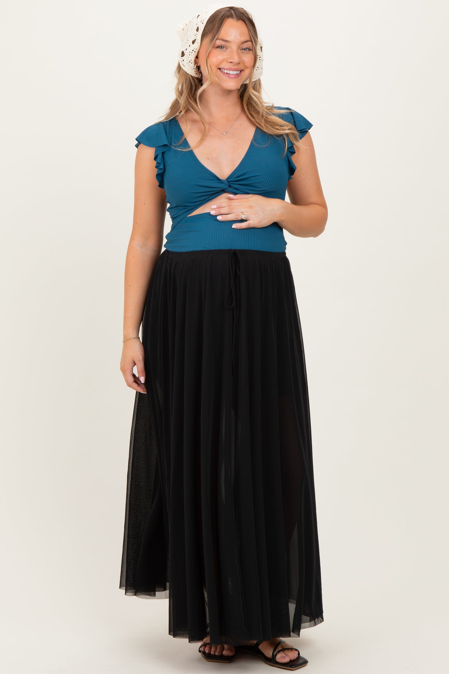 woman wearing a teal maternity top with ruffle sleeves and a black flowing skirt, holding her baby bump