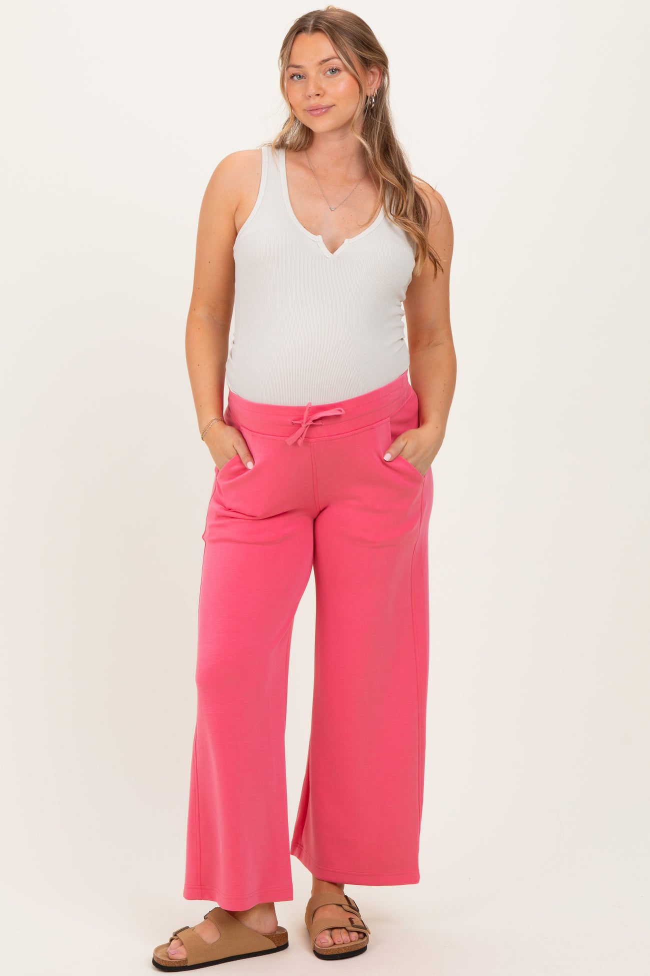 woman wearing a white tank top and pink maternity sweatpants with pockets and a drawstring waist, showing her baby bump
