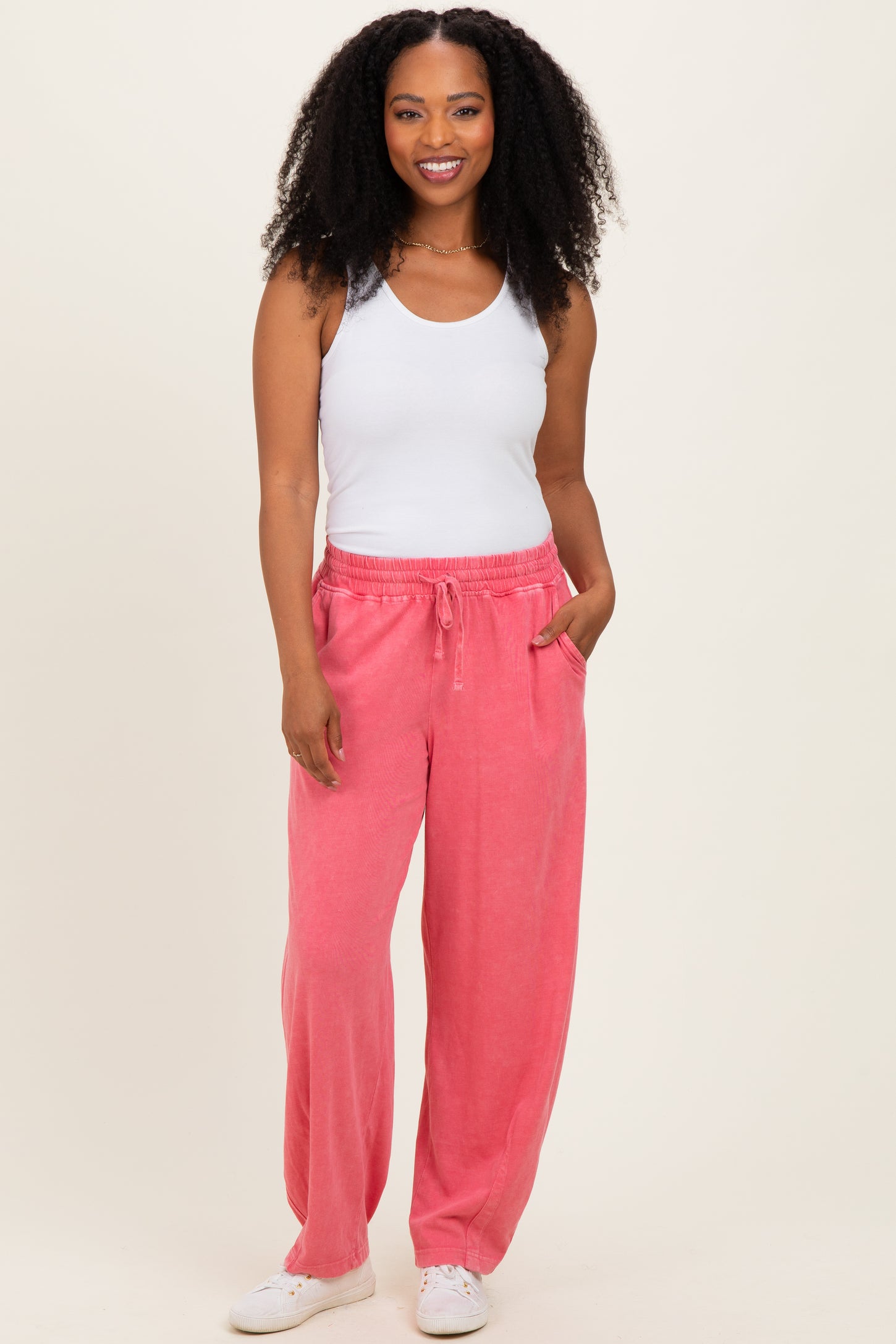 woman wearing a white tank top and pink relaxed fit pants with pockets, standing and smiling