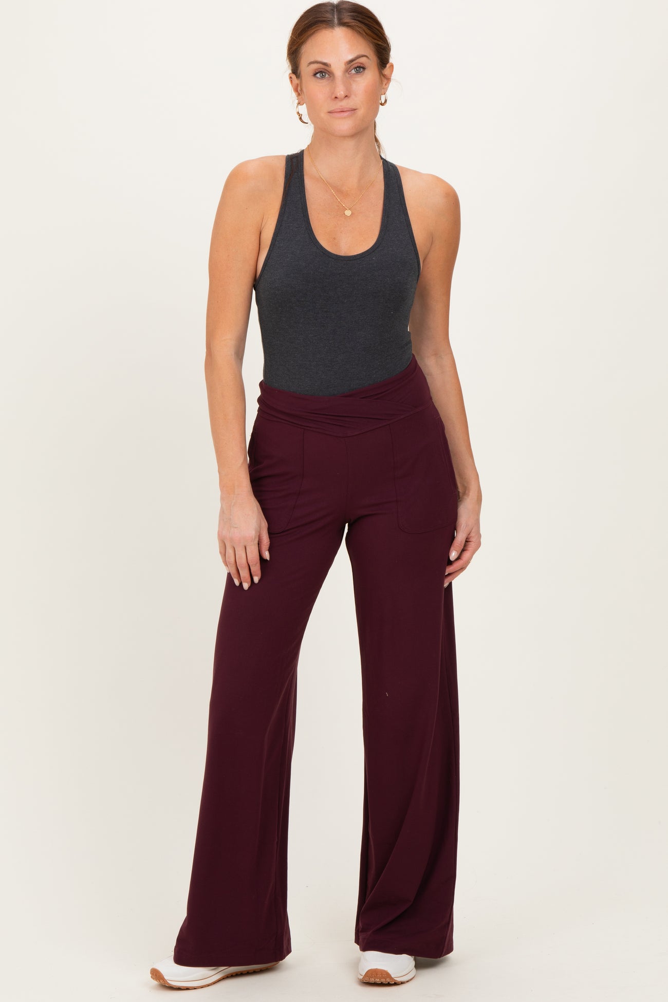 woman wearing a dark gray sleeveless top and burgundy wide-leg pants, standing with hands on hips against a light background