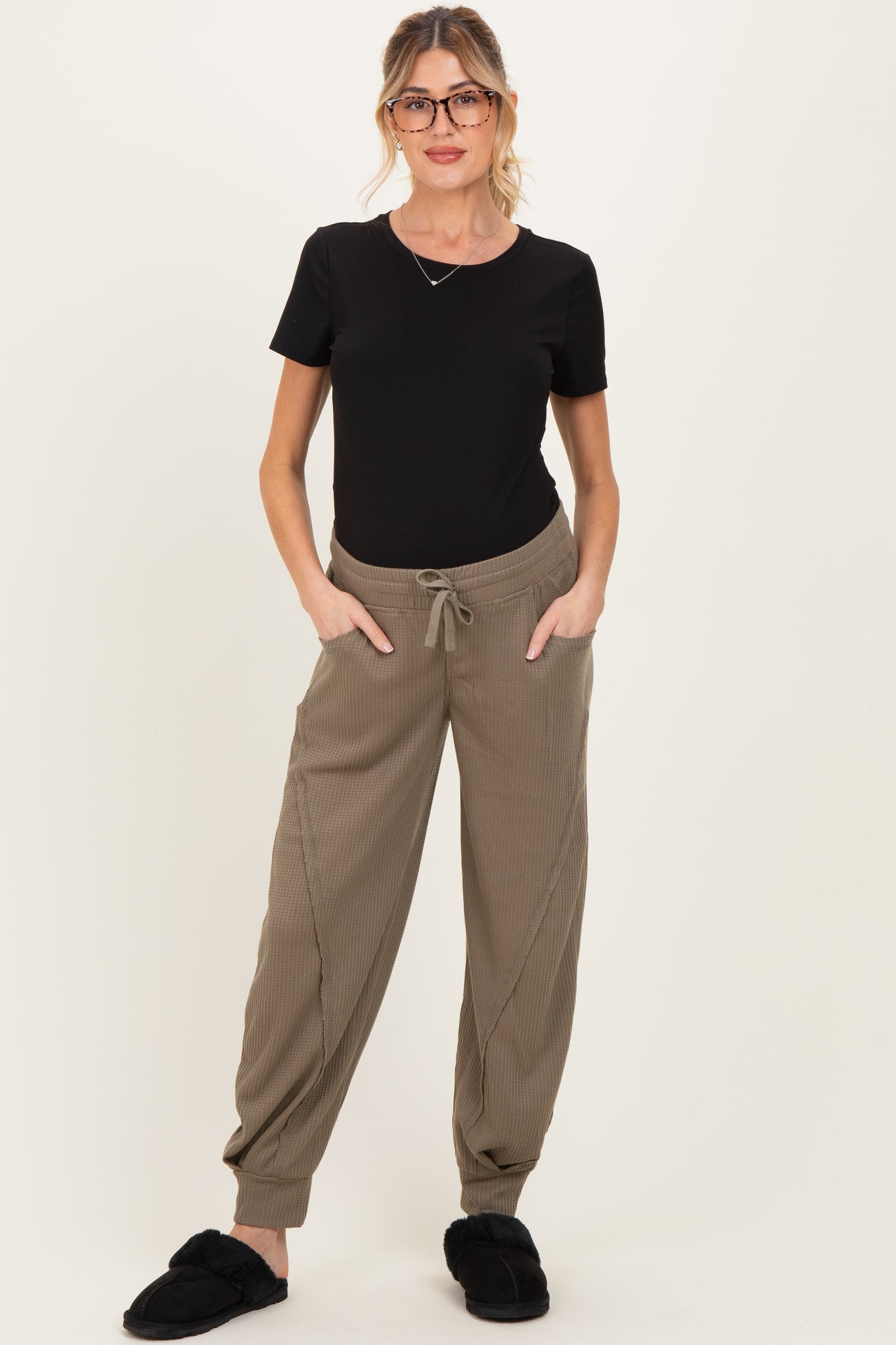 woman wearing a black maternity top and taupe ribbed maternity joggers with pockets, standing in cozy footwear