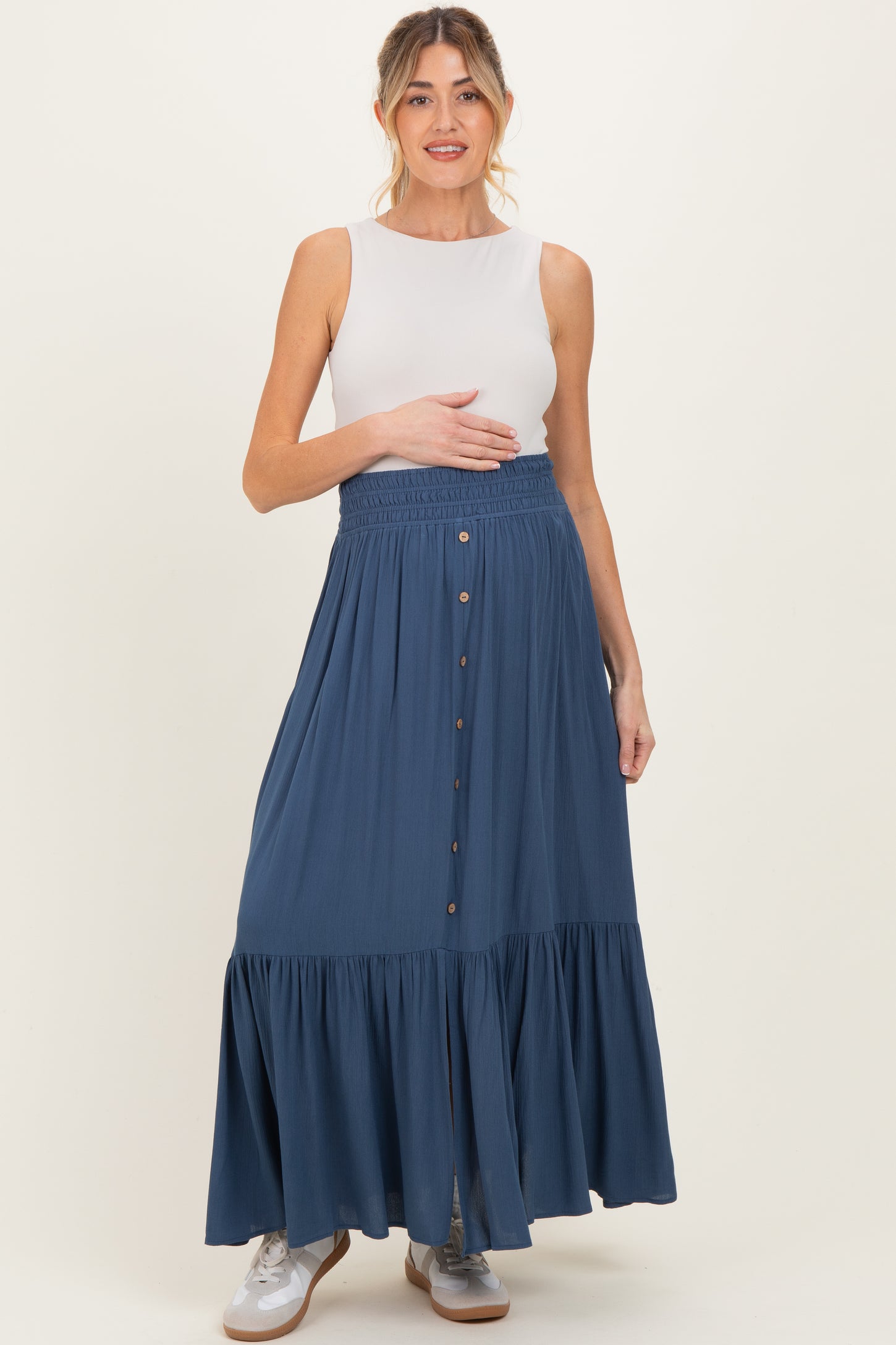 woman wearing a white sleeveless top and a long blue maternity skirt, holding her baby bump