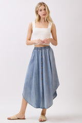 woman wearing a white ribbed tank top and a light denim button-up midi skirt, posing with a casual stance