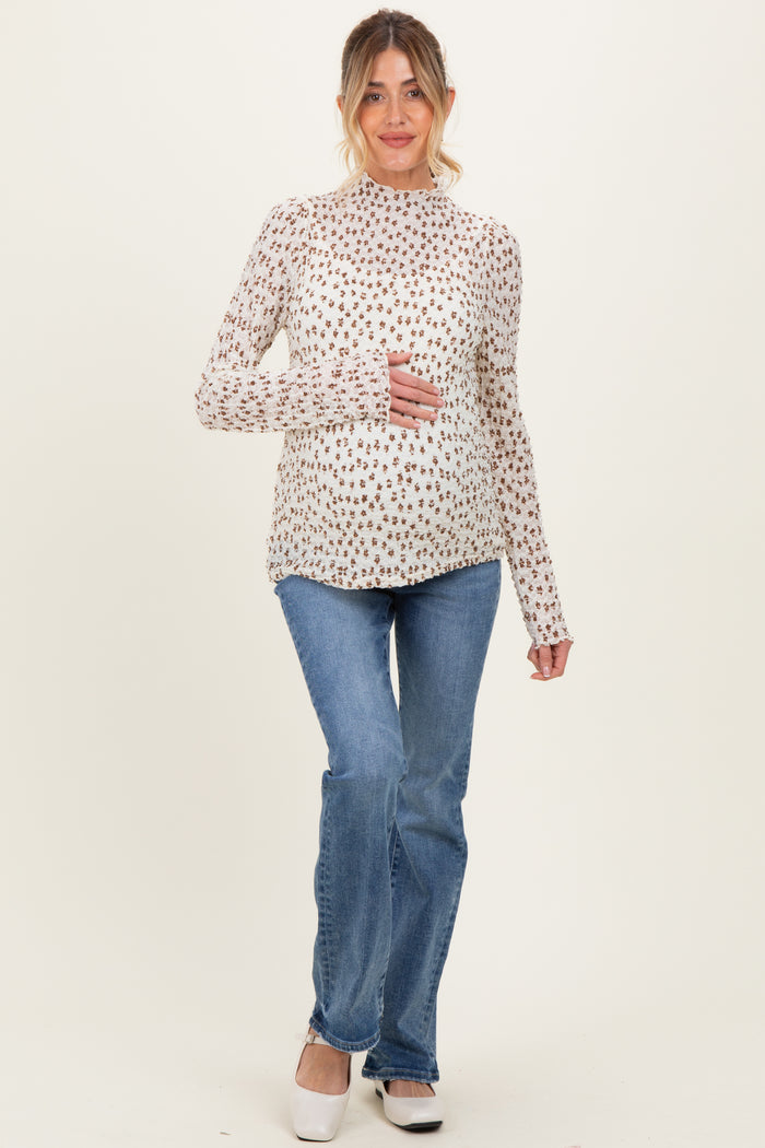 woman wearing a cream long sleeve maternity top with floral print and holding her baby bump, paired with blue flared jeans