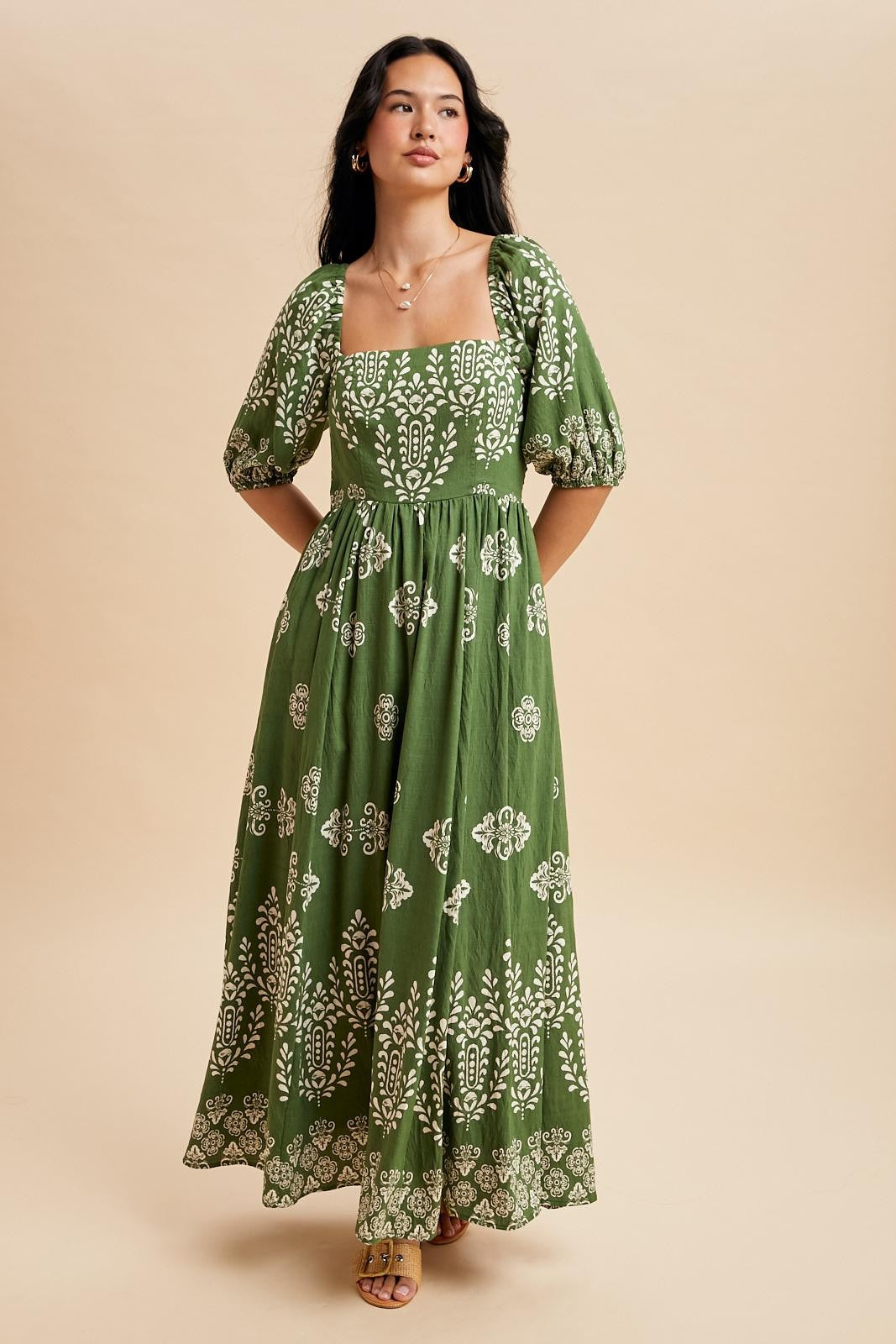 woman wearing a dark green maxi dress with puff sleeves and white floral patterns, standing against a light background