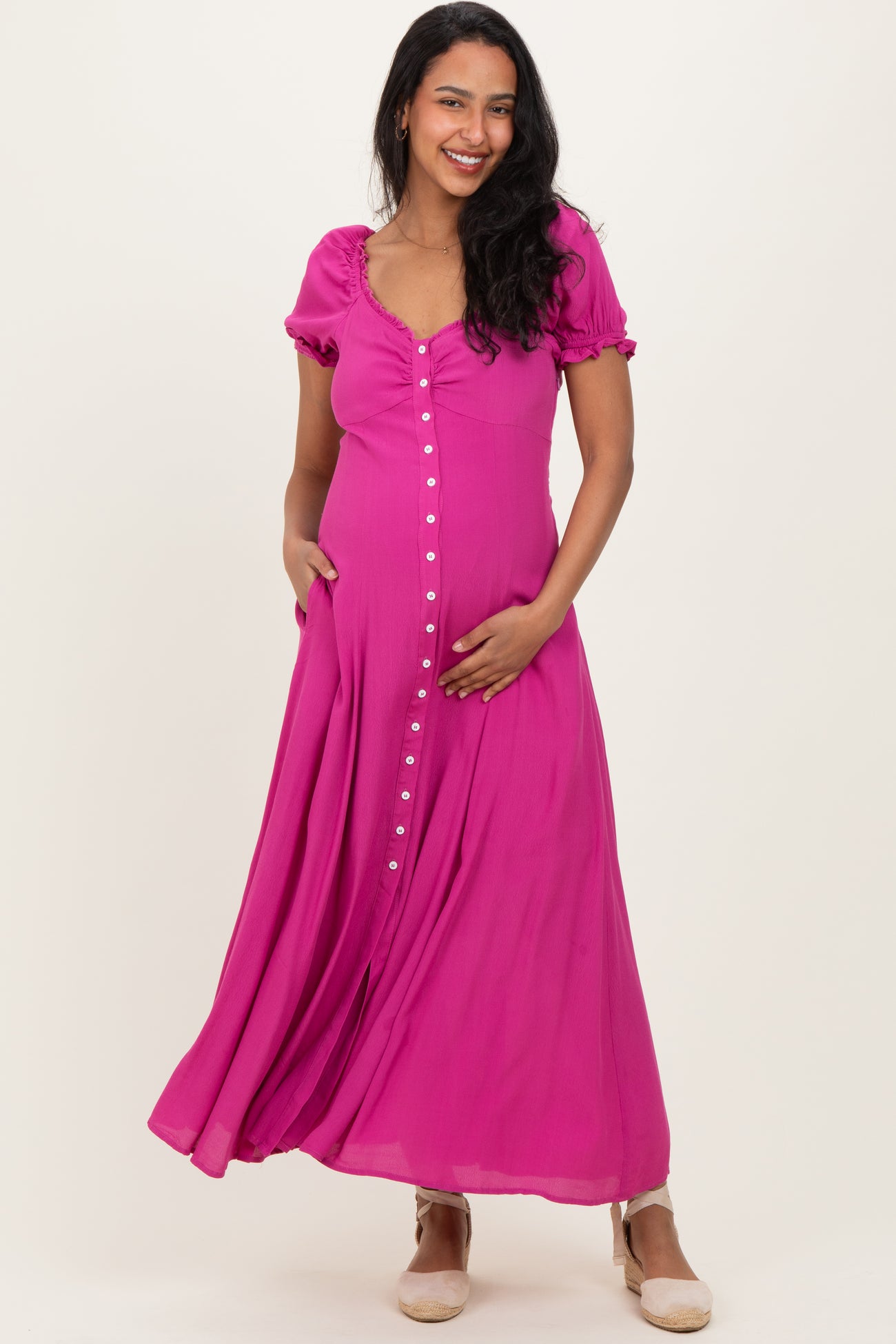 woman wearing a bright pink maternity maxi dress with short puff sleeves and button details, gently cradling her baby bump