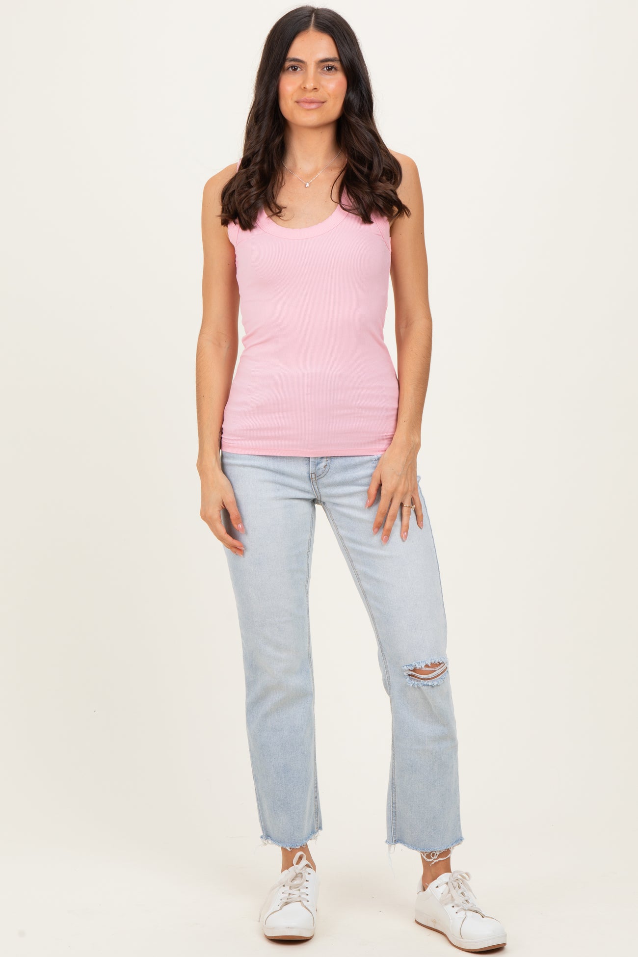 woman wearing a pink ribbed tank top and light blue ripped jeans, standing with a neutral background