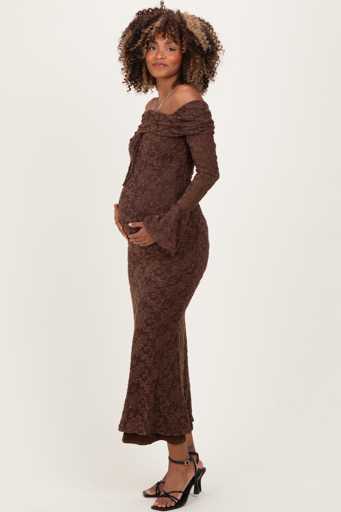 woman wearing a brown off-shoulder maternity dress with long sleeves and a fitted silhouette, cradling her baby bump