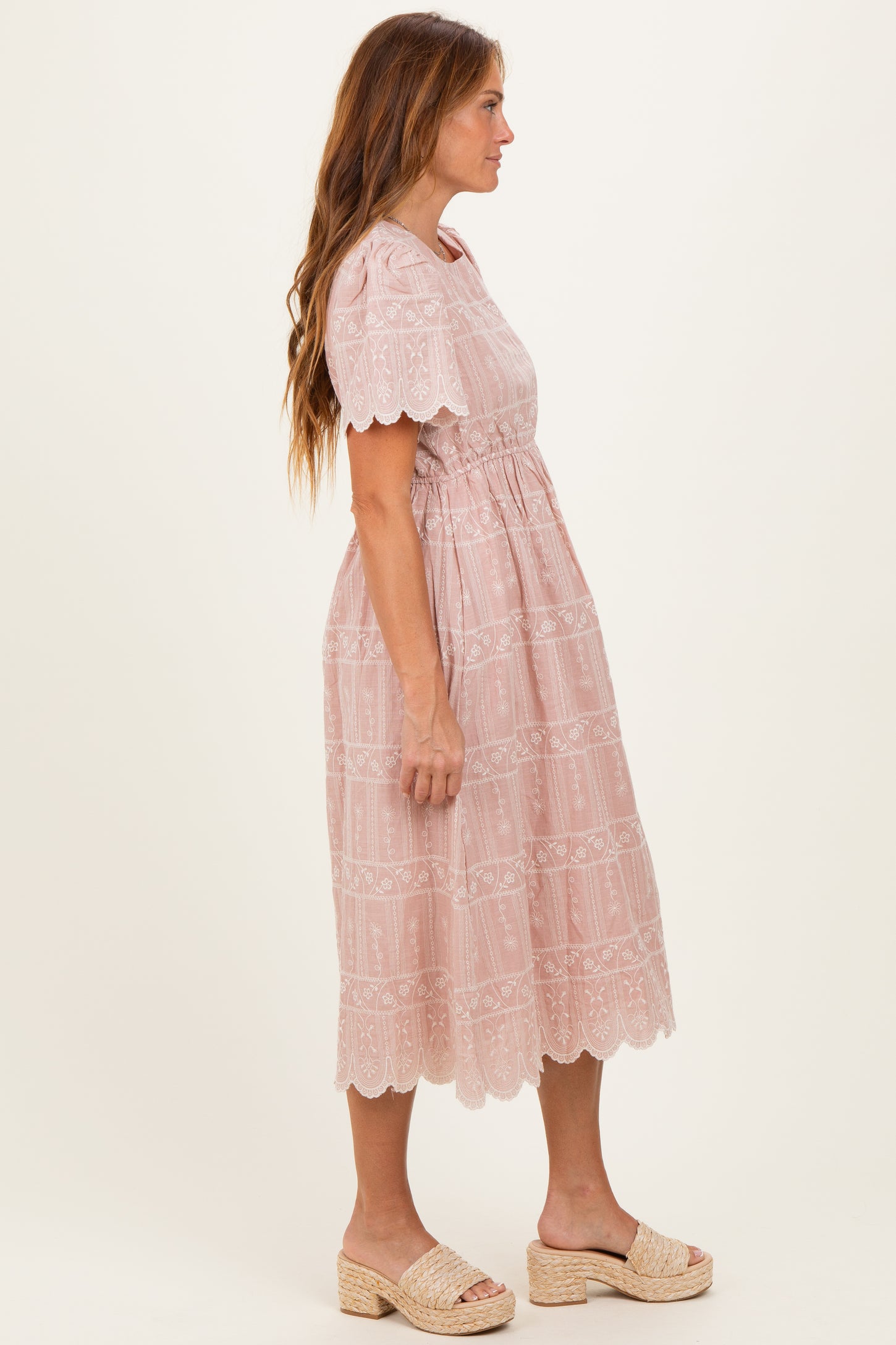 woman wearing a light pink scalloped edge midi dress with embroidered details and gathered waist, standing sideways