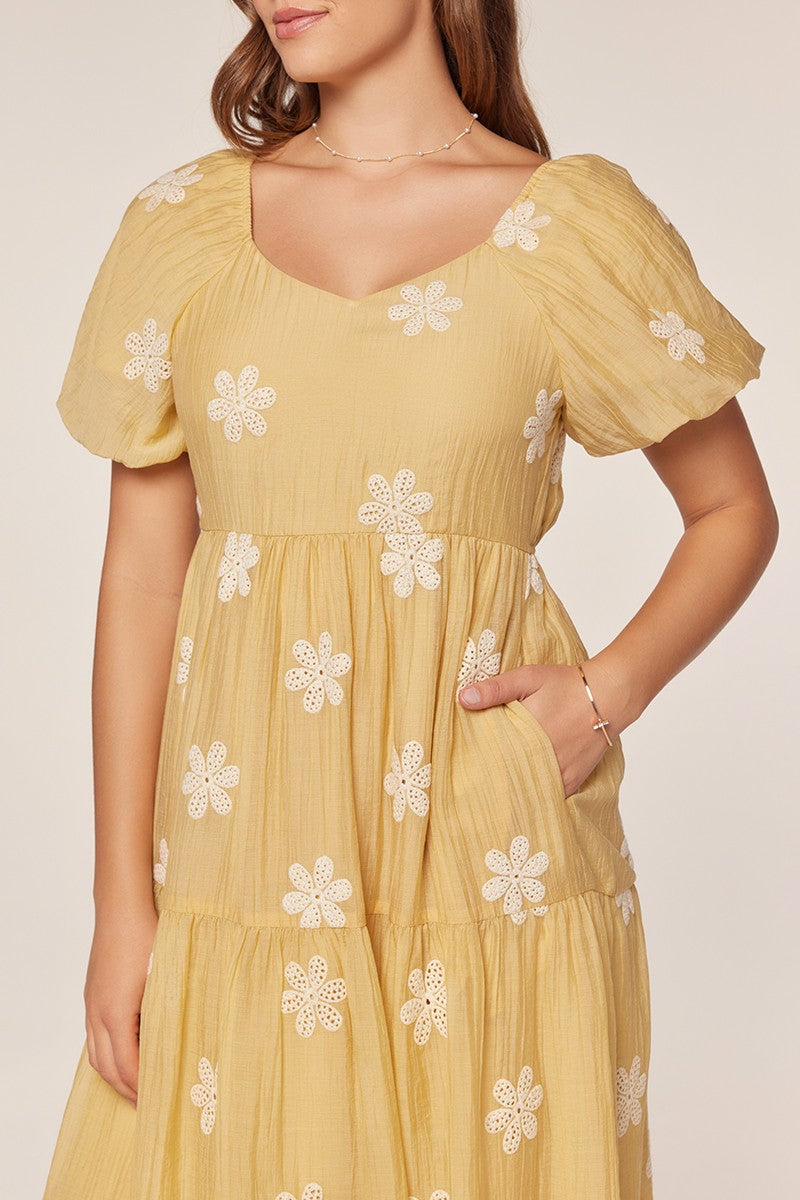 woman wearing a yellow tiered dress with puff sleeves and floral embroidery, featuring a square neckline and pockets