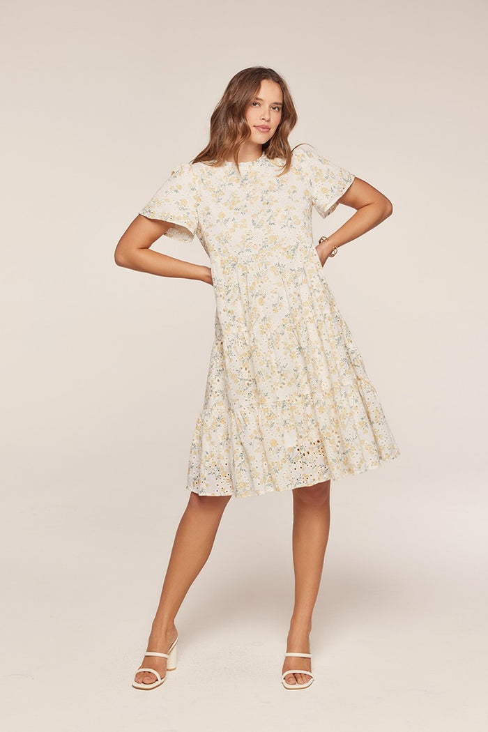 woman wearing a white floral short sleeve dress with a tiered skirt and eyelet details, standing with one hand on her hip