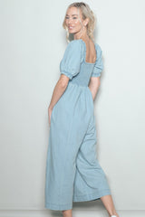 woman wearing a light blue denim jumpsuit with puff sleeves and a shirred back, smiling at the camera